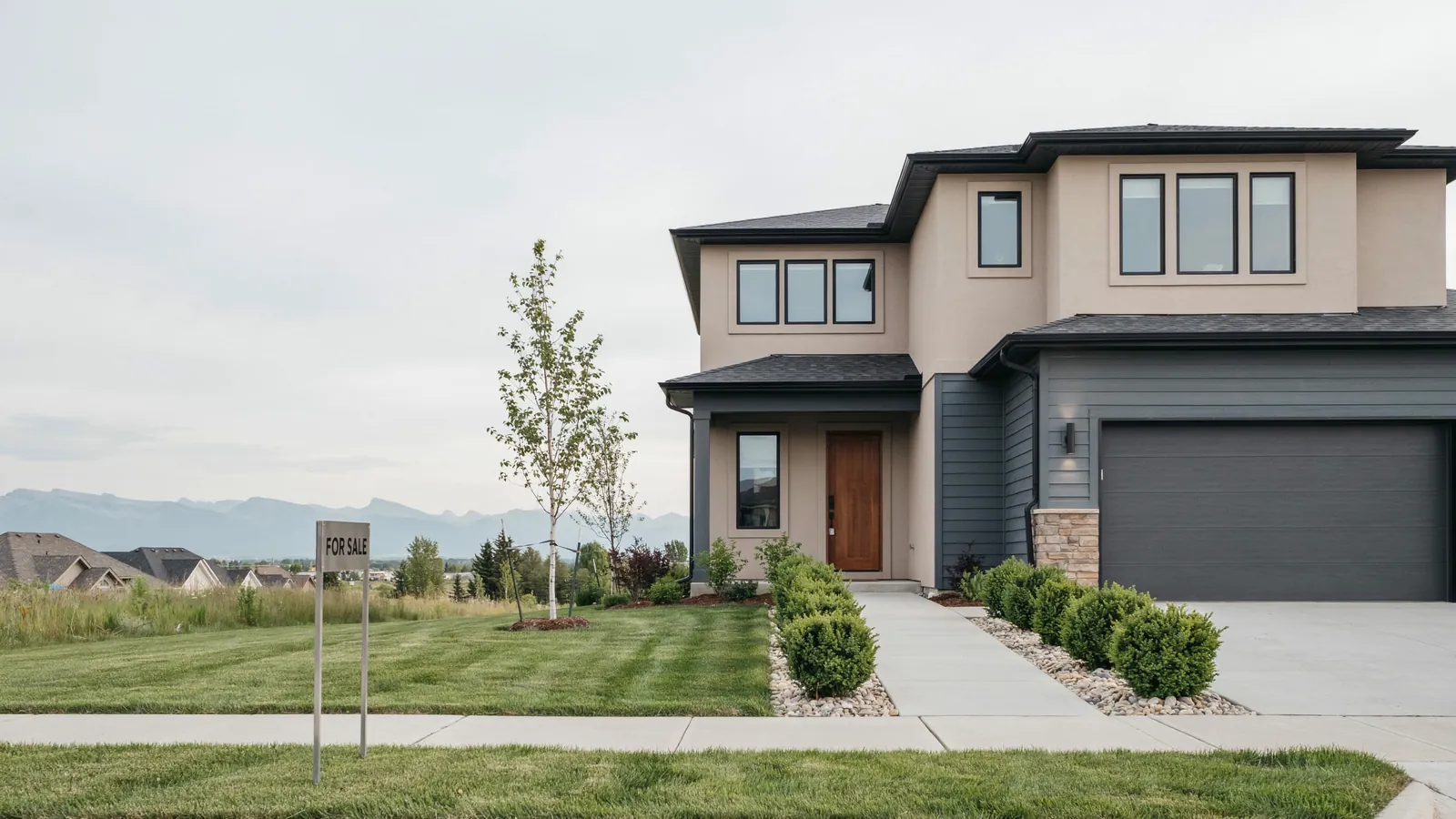 A modern two storey home with mountains behind it and a for sale sign on the lawn, the Alberta foreclosure guide